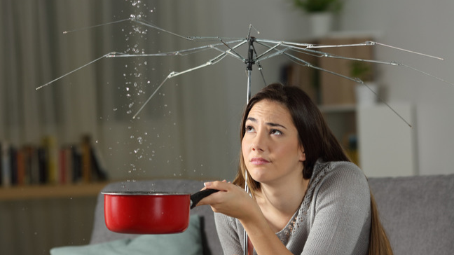 A frustrated woman holding up a broken umbrella and a pot, collecting water drippig from the ceiling.