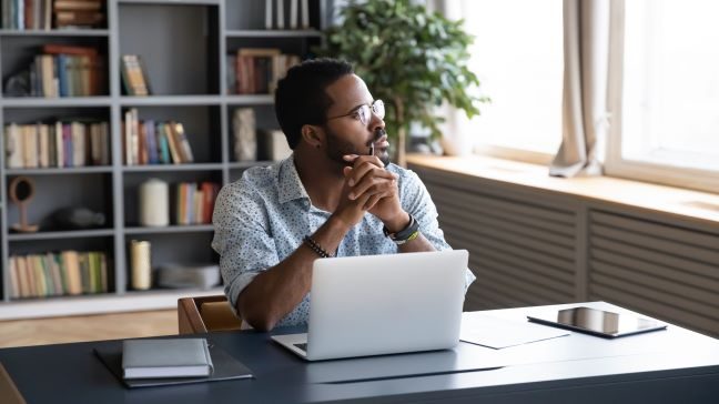 Person sitting in front of laptop and looking out the window with hands folded