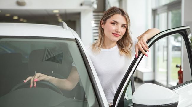 A young woman standing in the open driver's door of a new car, with one hand on the steering wheel.