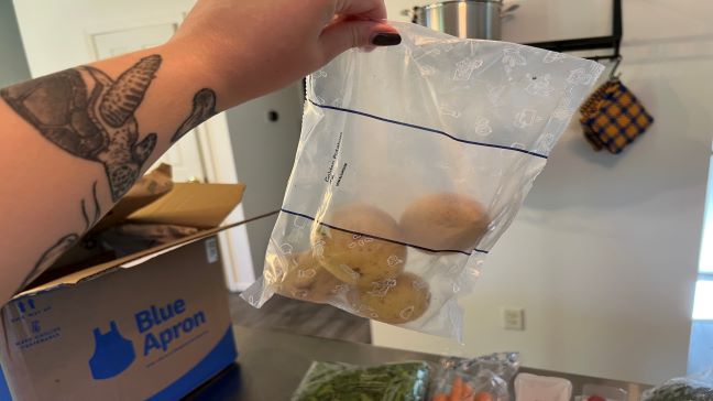 Holding up sealed plastic bag containing four small potatoes from Blue Apron meal kit