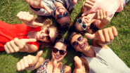 A group of friends wearing sunglasses and lying on the grass. Their heads are all together in a circle and they're giving the thumbs up.