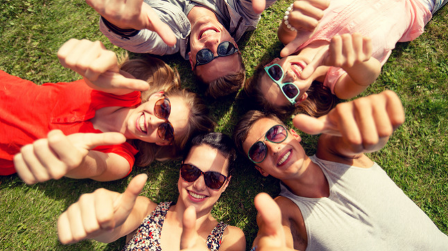 A group of friends wearing sunglasses and lying on the grass. Their heads are all together in a circle and they're giving the thumbs up.