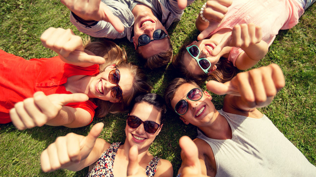 A group of friends wearing sunglasses and lying on the grass. Their heads are all together in a circle and they're giving the thumbs up.