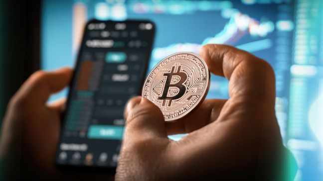 A hand holding a coin with a B on it, in front of a mobile app for investing