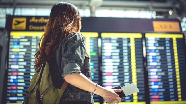 Young woman wearing a backpack, looking at the departures board in an airport, with a passport and ticket in her hand
