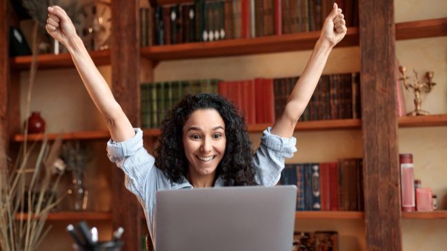 Woman throwing her arms in the air excitedely, while sitting in front of a laptop