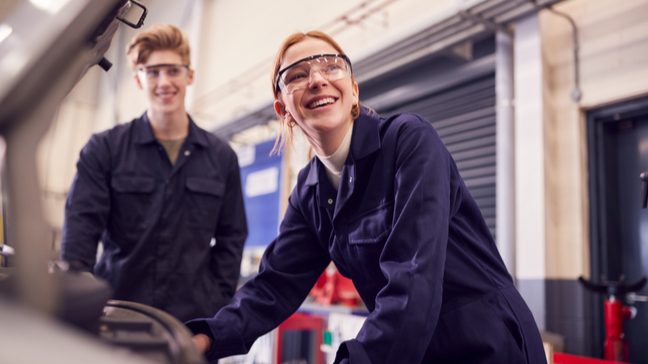 Two students in safety glasses and overalls, leaning over a machine