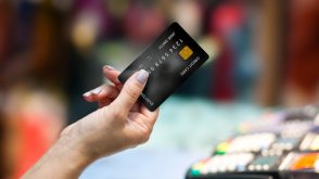 A woman's hand holding out a black credit card, with blurred store debit machines in the background.