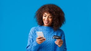 Smiling young woman in front of a blue background, looking at her phone with a credit card in her other hand.