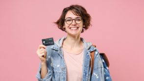 credit_card_teenager Smiling young woman student in denim clothes glasses backpack posing on a pastel pink background.