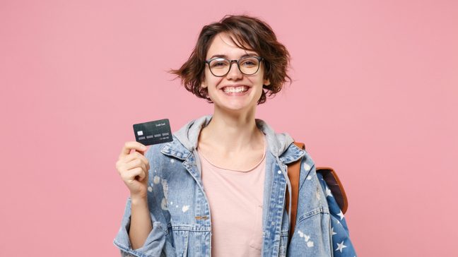 Smiling young woman student in denim clothes glasses backpack posing on a pastel pink background.
