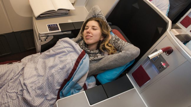 Young woman lying in a first class lie-flat seat on an airplane, with her eyes closed and arms stretched under her head