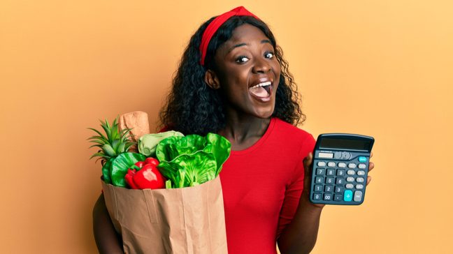 A woman holding a paper bag of fresh produce in one arm, and a calculator in her other hand.