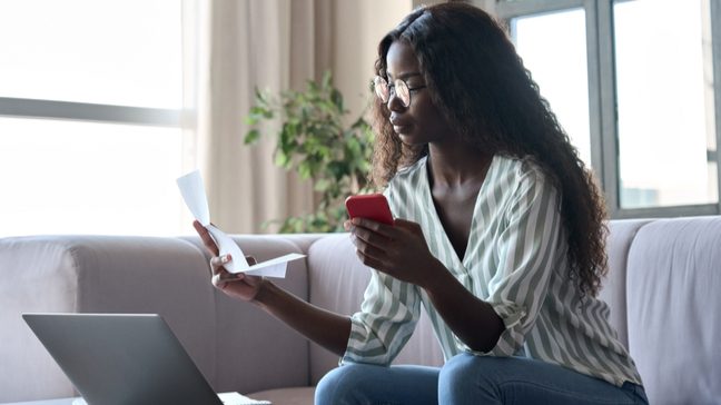 Young woman sitting on a couch, looking at her phone, a laptop, and paperwork