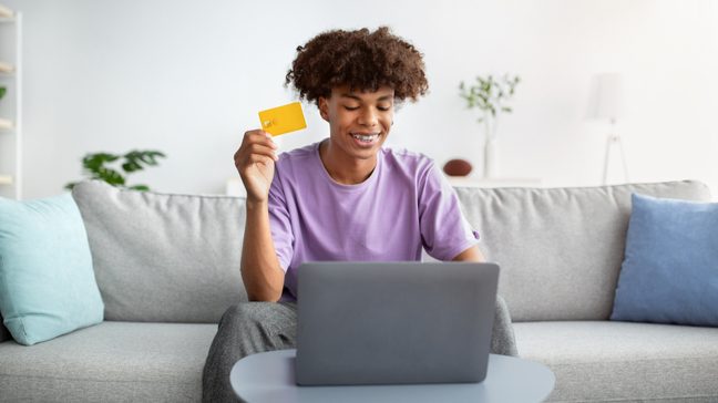 Teenager sitting on a couch, in front of a laptop and holding a credit card