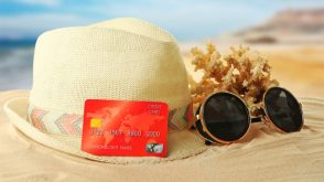 A red credit card propped up against a sun hat on a sandy beach, with sunglasses beside it.