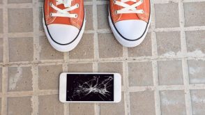A cellphone with a broken screen lying on the ground, in front of a pair of feet wearing sneakers.