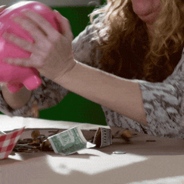 A gif of a woman shaking a pink piggy bank, with a small pile of coins and dollar bills on the table in front of her.