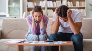 A white man and woman sitting on a living room couch side-by-side, each leaning over a coffee table scattered with paperwork. Both look frustrated, with their heads in their hands.