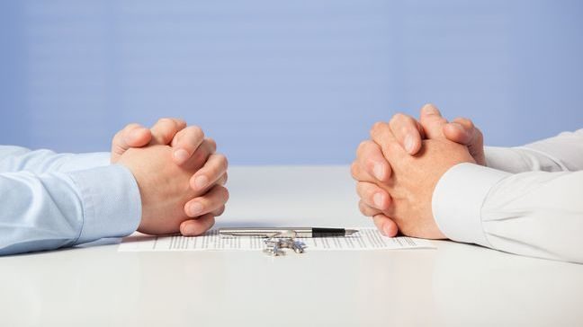 Two sets of hands, clasped and resting on a table with a contract, pen, and keys in between them.