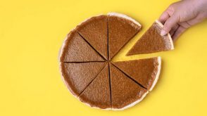 An overview shot of a pumpkin pie, cut into 8 pieces, with a hand taking one piece.