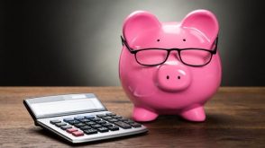 A pink piggy bank wearing glasses, next to a calculator on a wooden table.