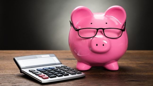 A pink piggy bank wearing glasses, next to a calculator on a wooden table.