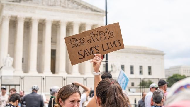 A hand holding up a cardboard sign in a crowd of protestors, that reads 