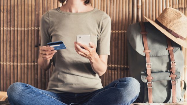 Young woman sitting cross-legged, holding a cellphone and credit card, with a travel backpack beside her.