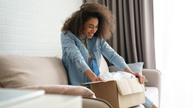 A smiling woman sitting on the couch and opening a brown box delivery.