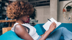 A young Black woman reclining on a couch and writing in a journal.