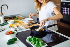 Woman cooks at home using tongs in a pan with vegetables off to the side