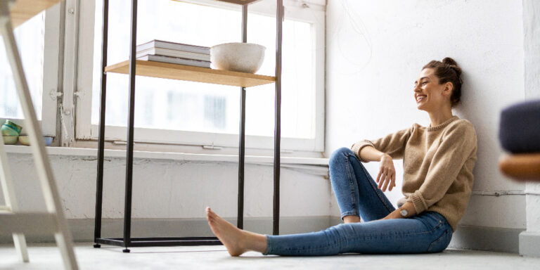 A young woman sitting on her apartment floor looking happy. From "The Cost of Living Alone".