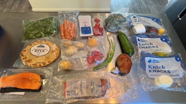 Ingredients from a Blue Apron meal kit spread out over kitchen island with some in plastic packaging, including pita, salmon, carrots, potatoes and green onions
